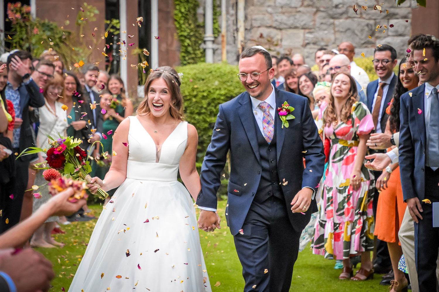 Joyful bride and groom walk through colorful confetti shower surrounded by celebrating wedding guests.