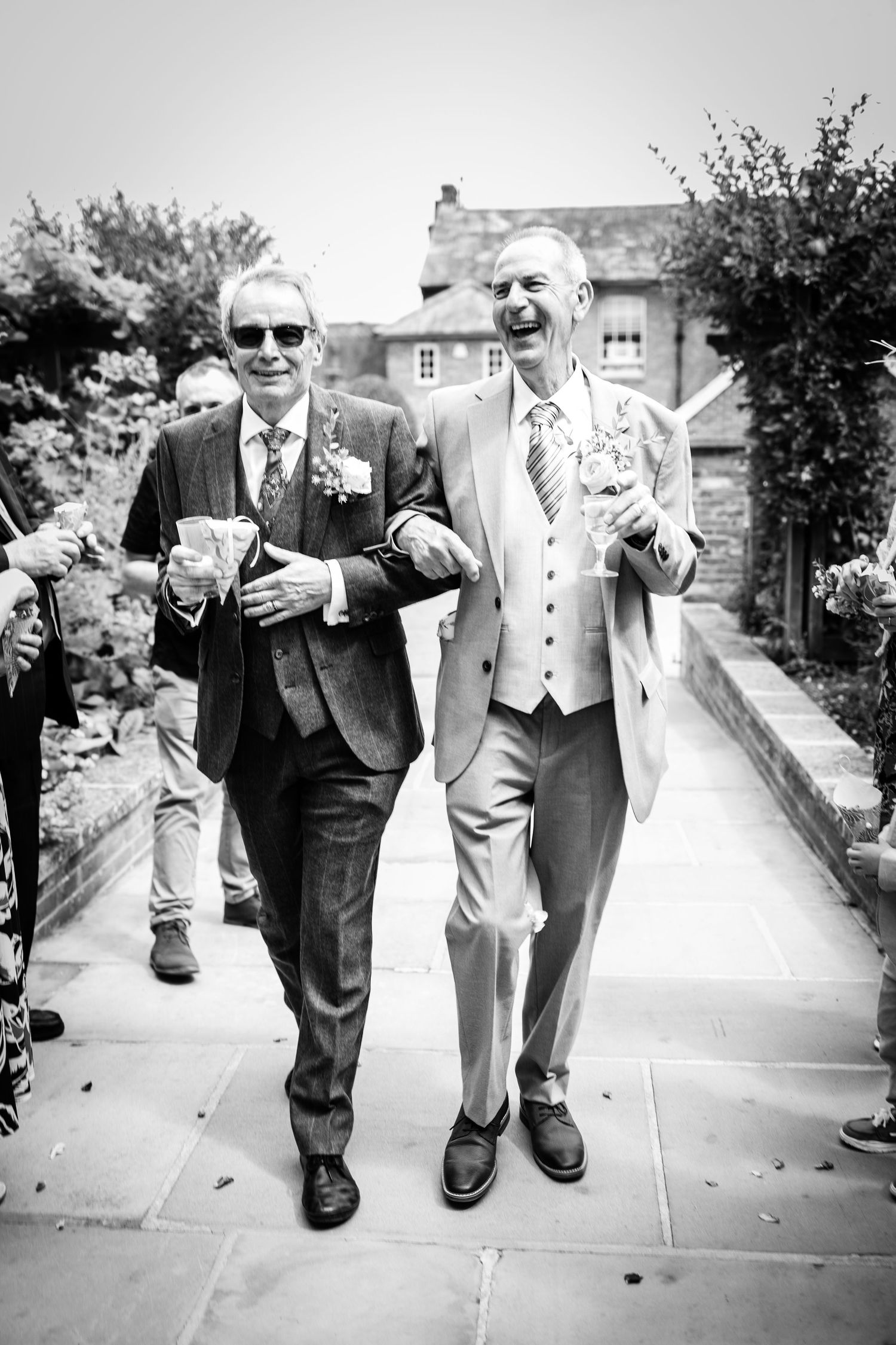 Two grooms walk arm-in-arm smiling at their wedding celebration in black and white photography.