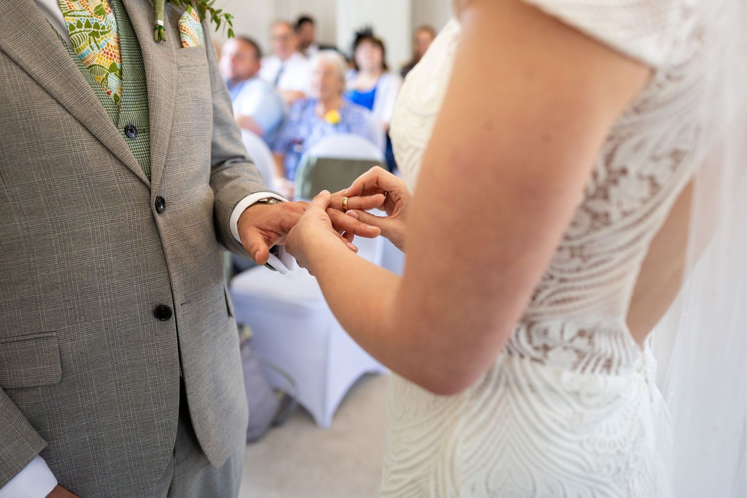 Bride placing wedding ring on groom's finger during intimate ceremony with guests seated in background.