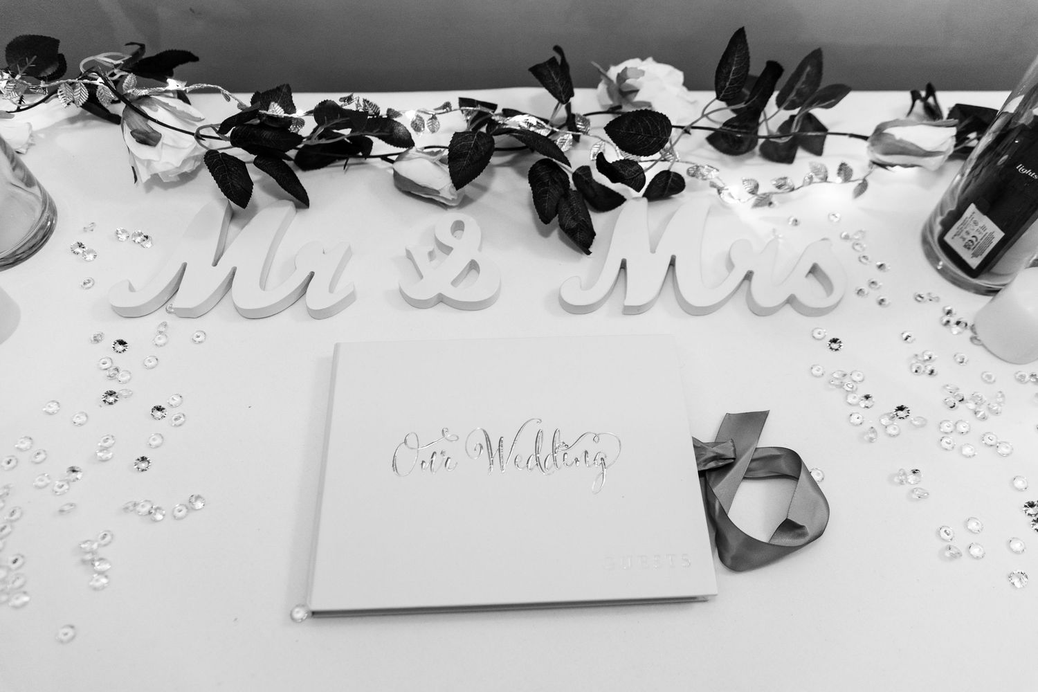A black and white wedding photo showing a guest book with 'Mr & Mrs' signage and scattered decorative elements on a table.