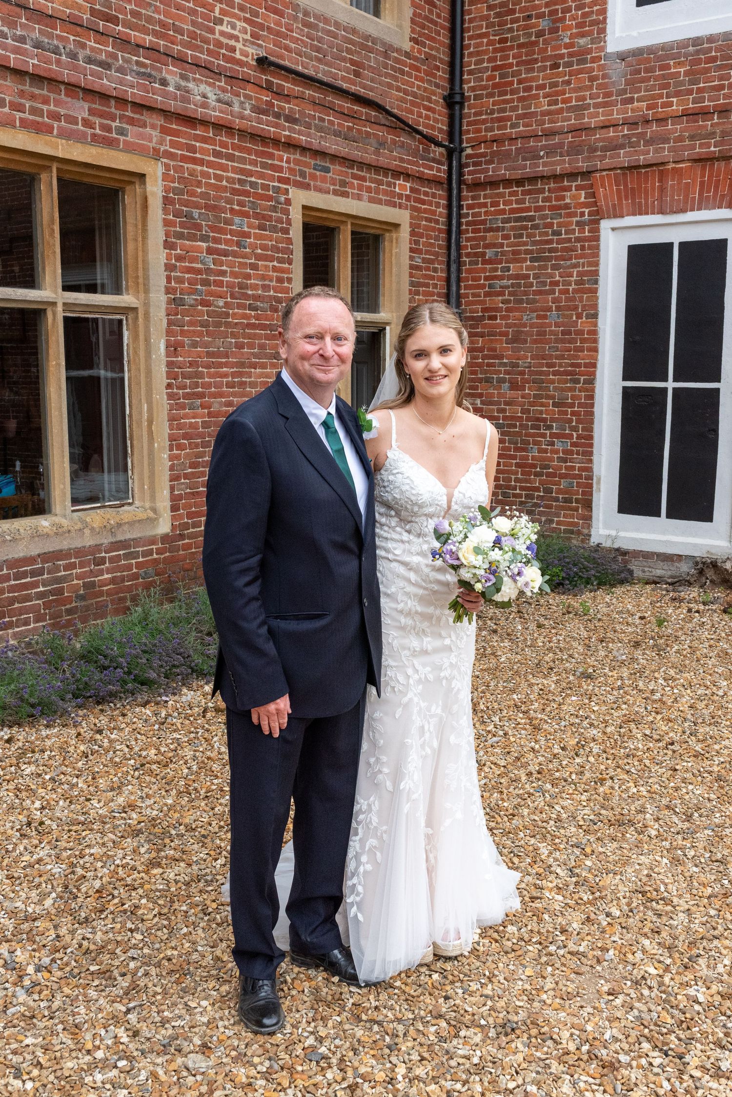 Bride holding floral bouquet in lace wedding gown poses with suited man outside brick building.
