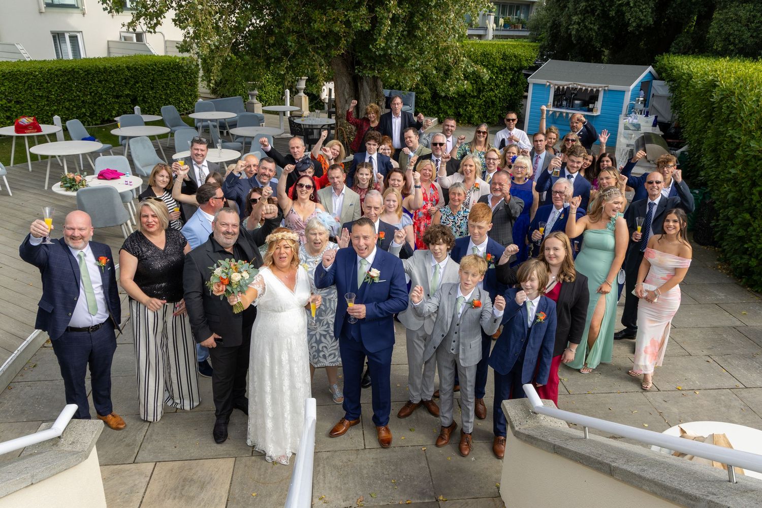 Large wedding group photo outdoors with bride and groom at center, surrounded by smiling guests raising glasses.