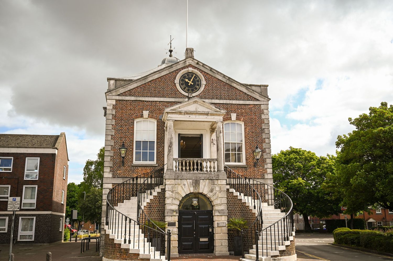 Historic Georgian brick town hall with clock tower, grand stone staircase, and black iron railings under cloudy sky.