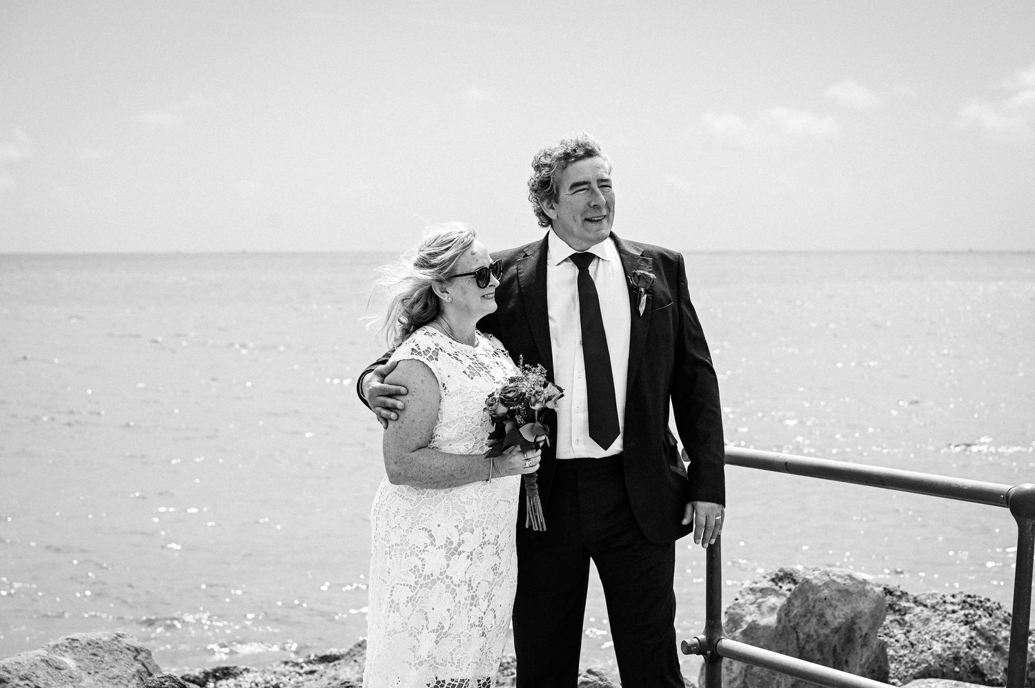 Black and white photo of a bride and groom standing by the sea, bride holding a bouquet, both looking into the distance.