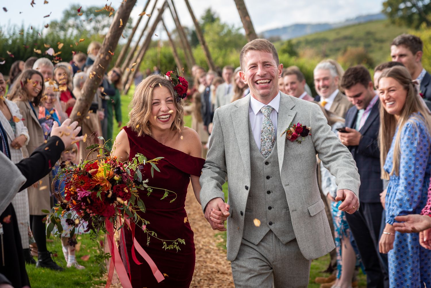 Joyful bride in burgundy dress and groom in gray suit walk through confetti shower at outdoor wedding ceremony.