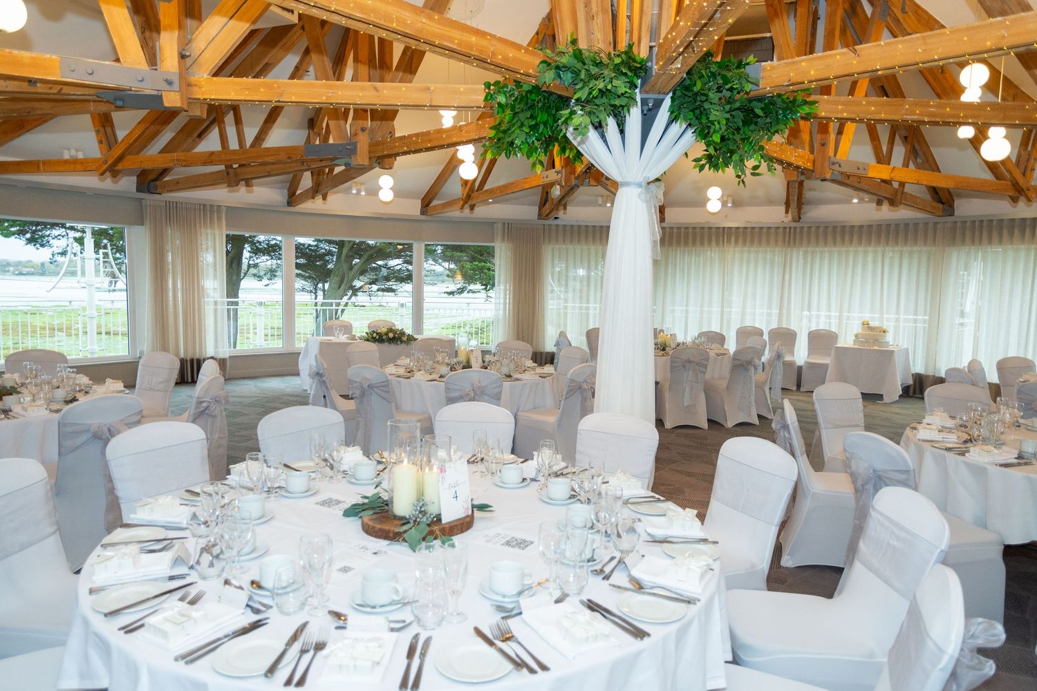Elegant wedding reception hall with white chair covers, floral centerpieces, and wooden beam ceiling with garden views.