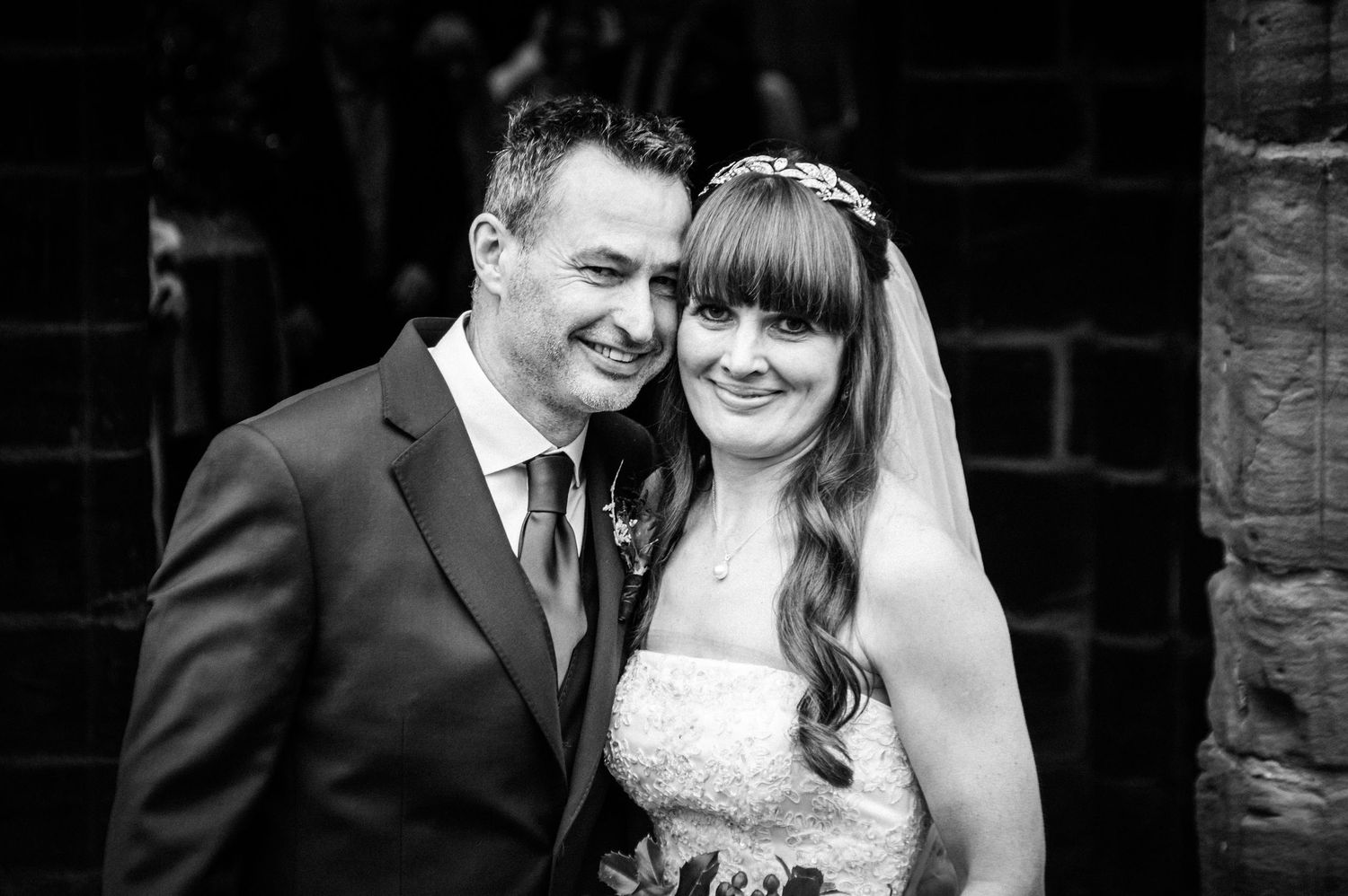 Black and white wedding photo of smiling bride and groom posing together outdoors near a brick wall.
