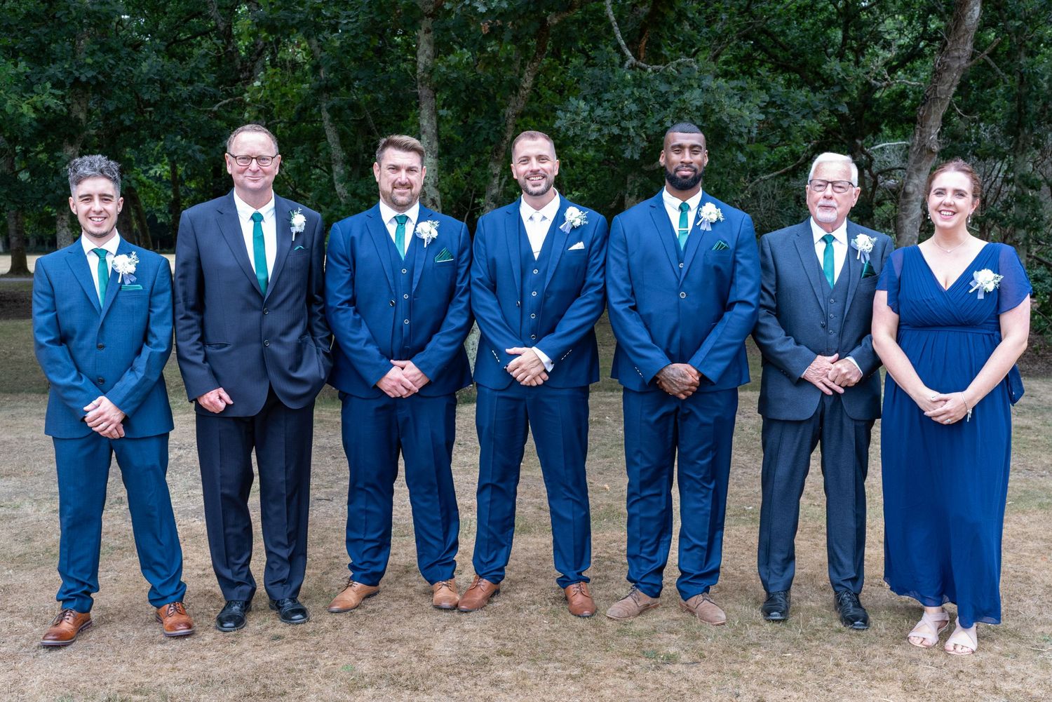 Seven wedding party members in navy blue suits and dresses posing outdoors for a formal group portrait.