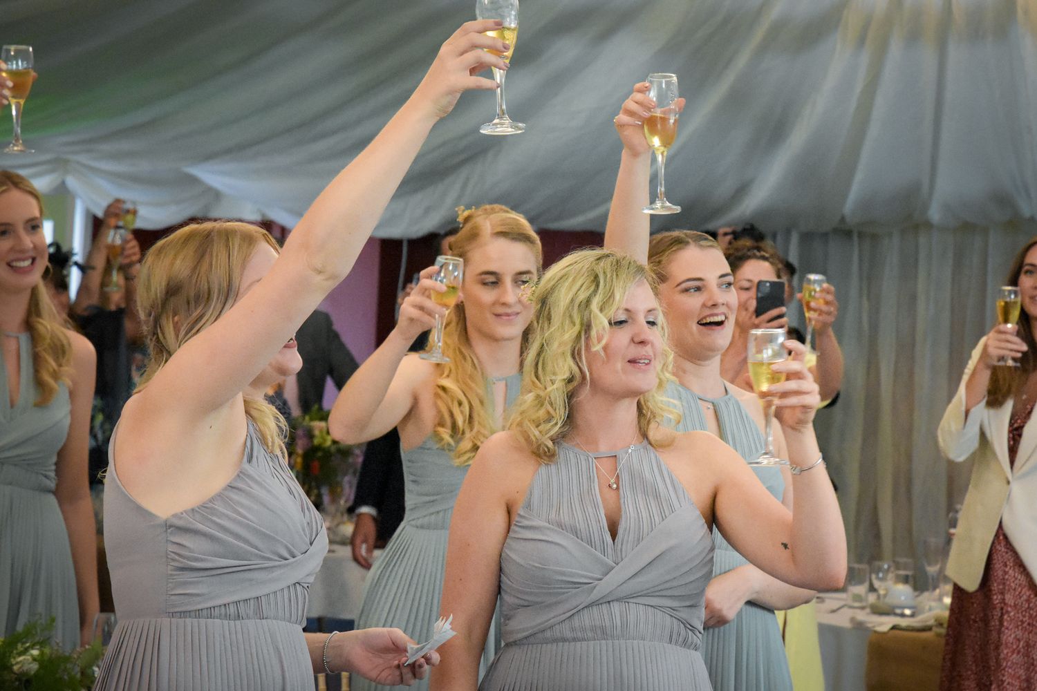 Bridesmaids in grey dresses raise champagne glasses in a celebratory toast at a wedding reception.