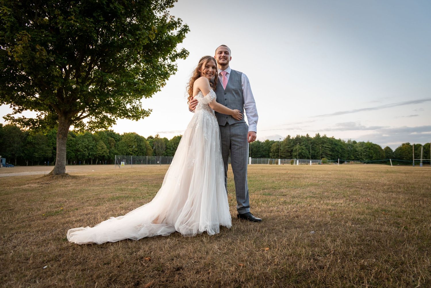 Bride and groom pose together on open field at golden hour, bride in elegant white gown with long train.