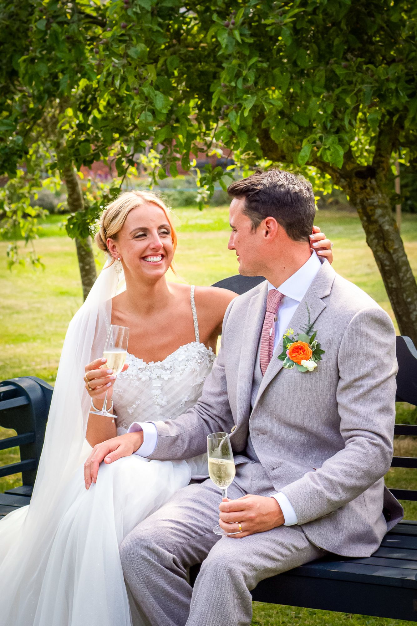 Bride and groom sitting on a bench, smiling and holding champagne glasses, surrounded by lush green trees.