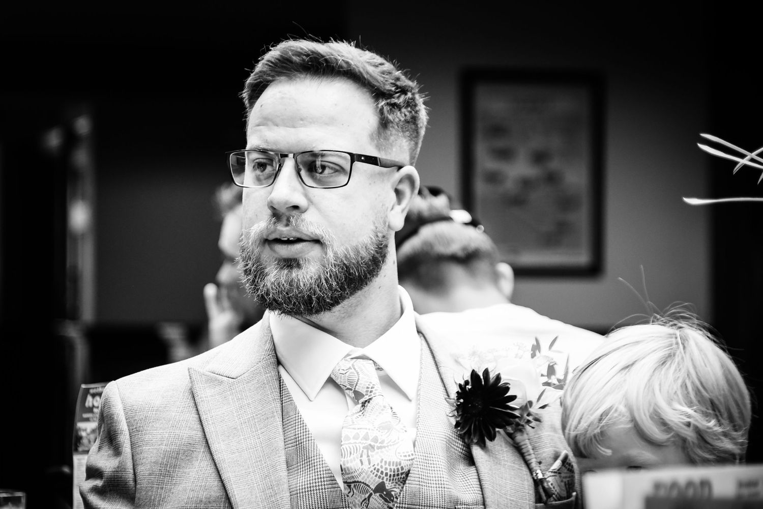 Black and white photo of a bearded groom wearing glasses and a floral boutonniere at a wedding ceremony.