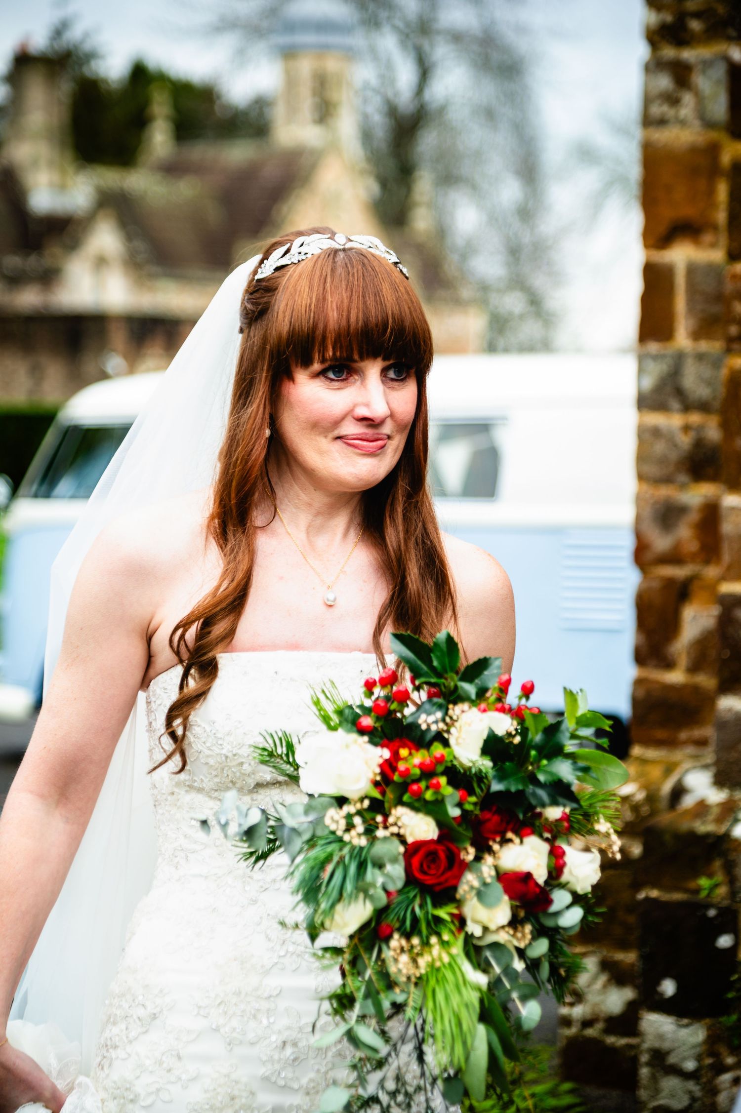 A bride in a white lace dress holds a vibrant red and green bouquet with eucalyptus leaves against a stone wall backdrop.