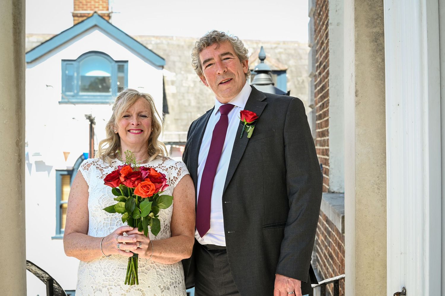Smiling couple posing outdoors on their wedding day, bride holding red roses bouquet, groom wearing dark suit with red rose boutonniere.