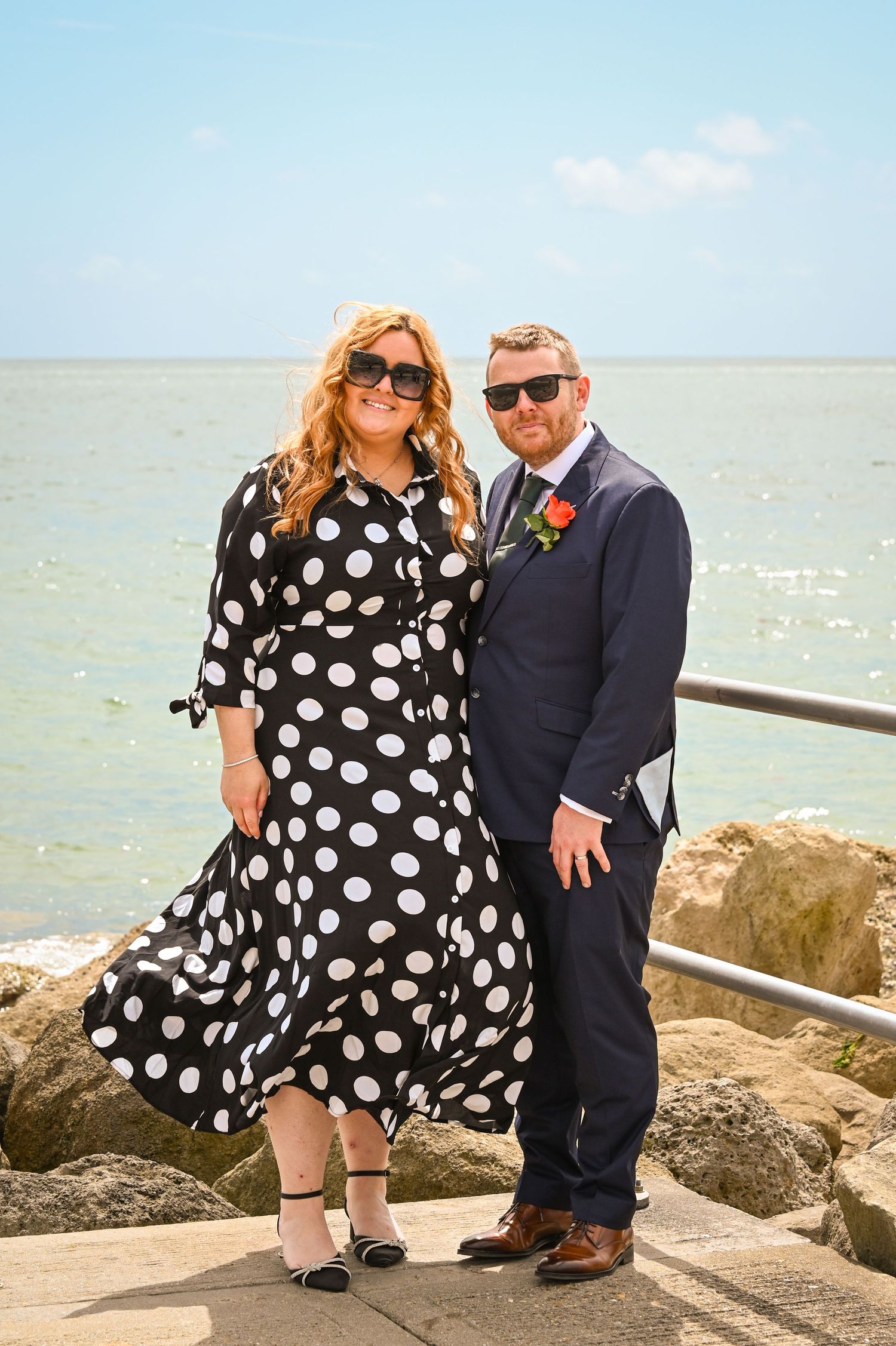 Couple in formal attire posing by the seaside, woman in polka dot dress and man in navy suit with boutonniere.