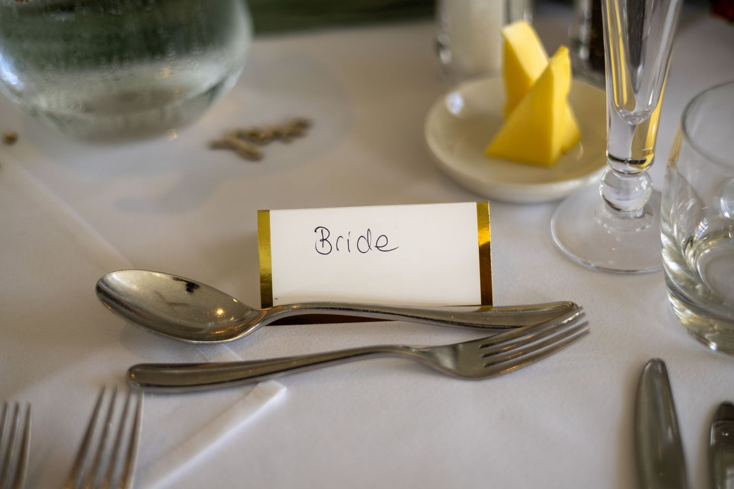 Elegant wedding table setting with a gold-trimmed Bride place card, silverware, and yellow napkin on white linen.