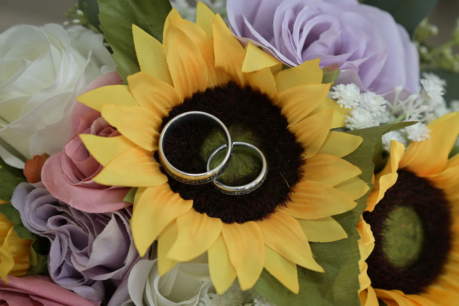 Two silver wedding rings resting on a yellow sunflower surrounded by colorful roses in a bridal bouquet.
