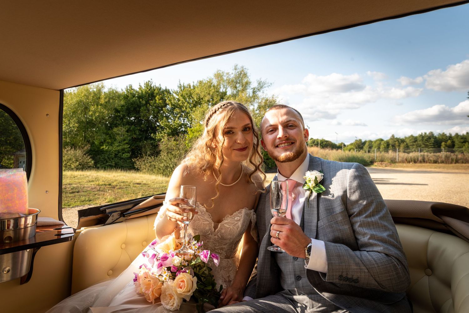 Bride and groom sitting in a vintage car, holding champagne glasses and a bouquet on their wedding day.