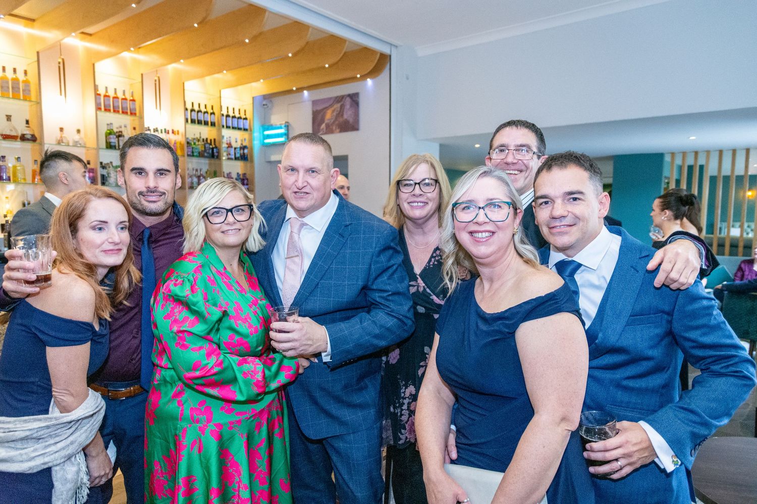 Group of smiling guests posing together at a formal party event in a modern bar venue.