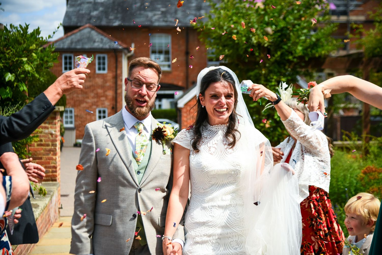 Bride and groom walking through colorful confetti shower outside brick venue on sunny wedding day.