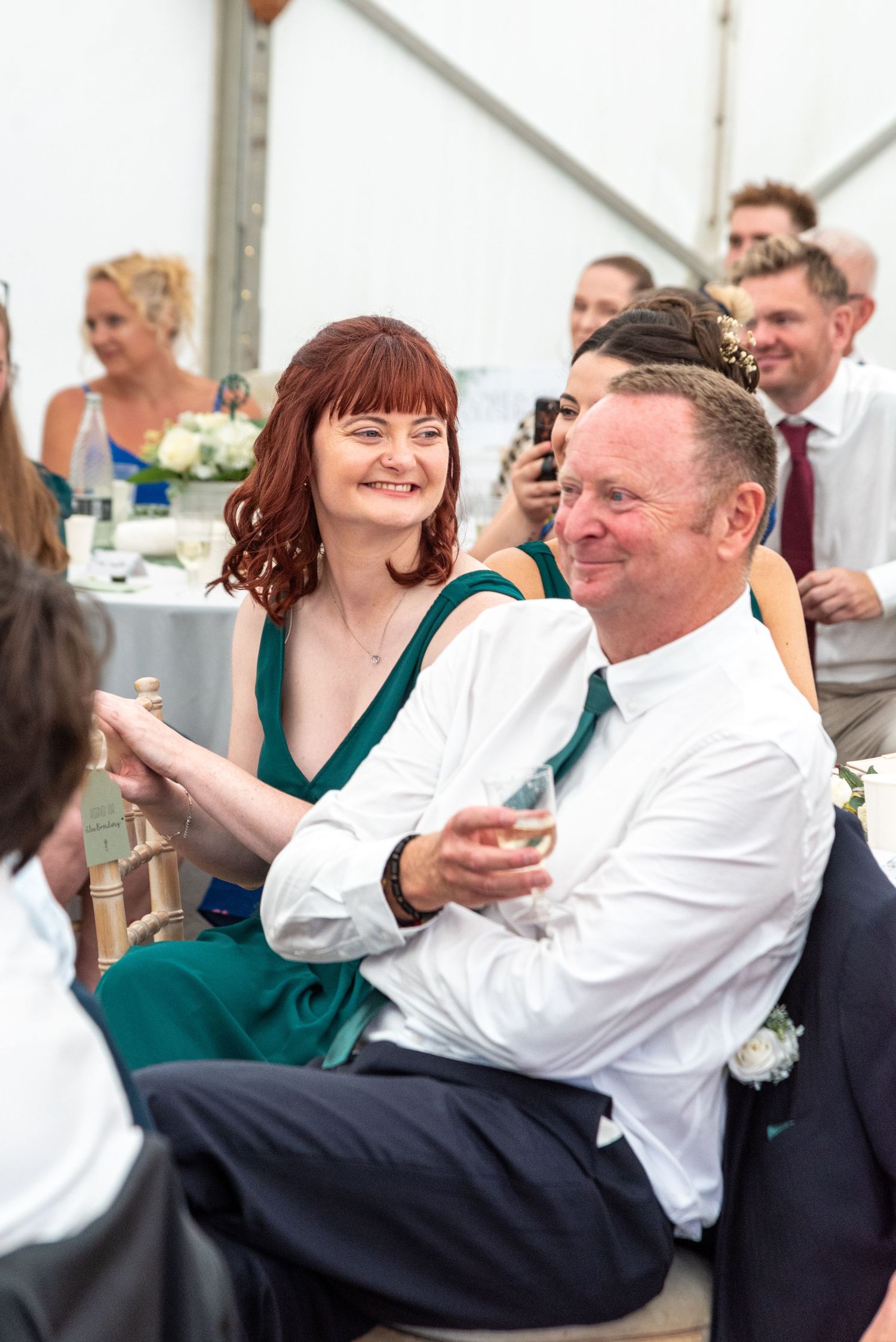 Wedding guests in emerald green and navy laughing together at a reception under a white marquee tent.