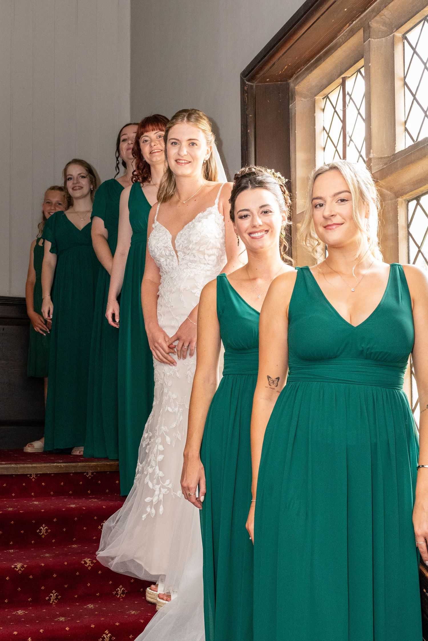 Bride in white lace gown poses with bridesmaids in emerald green dresses on red-carpeted stairs.