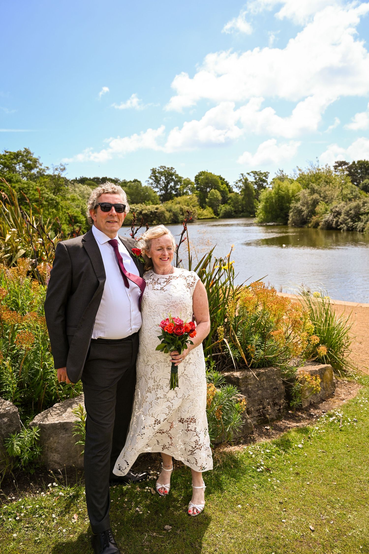 Bride and groom pose by a lakeside garden, bride holding red bouquet in white lace dress, groom in dark suit with sunglasses.