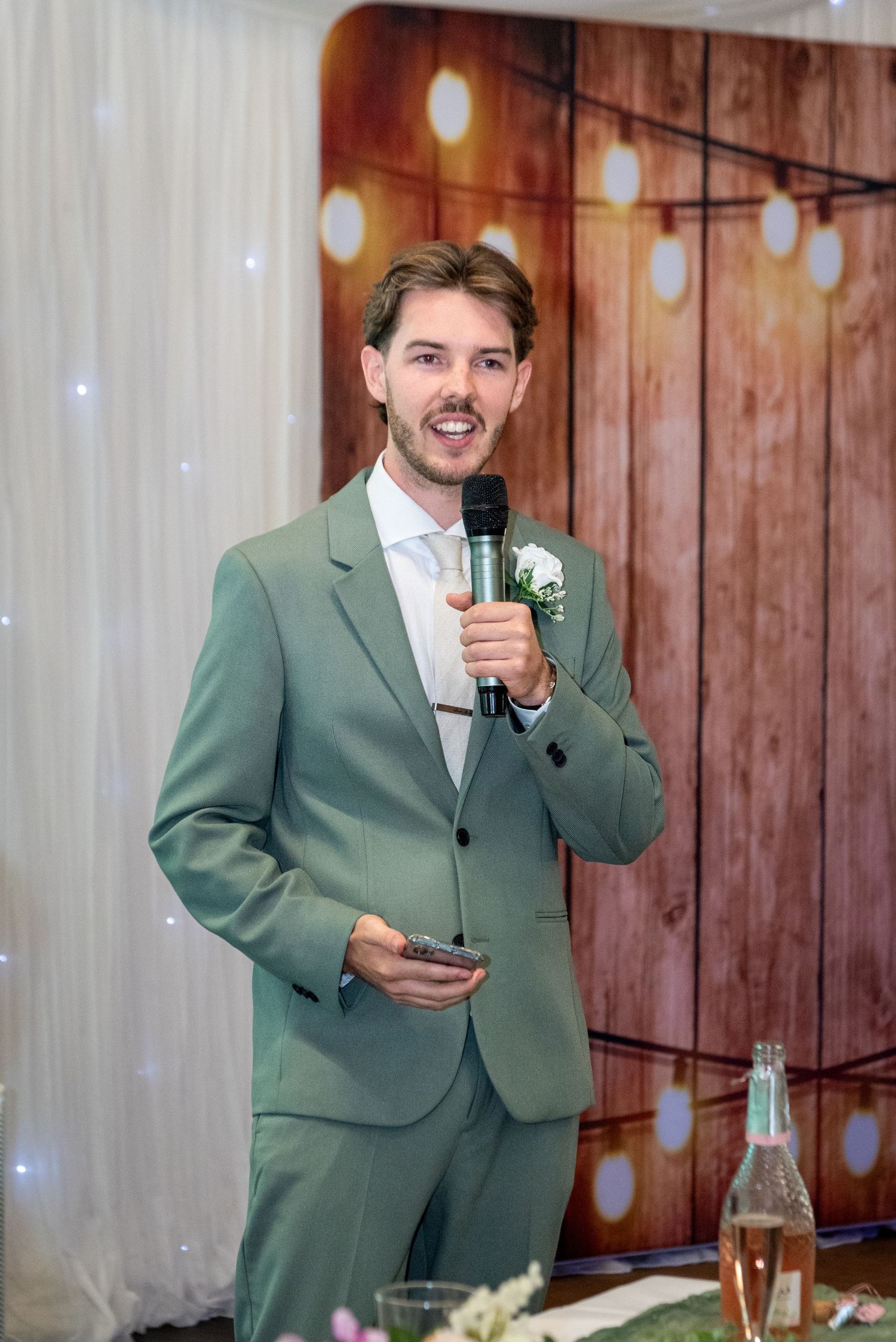 Groom in sage green suit giving a wedding speech while holding a microphone and phone at a reception.
