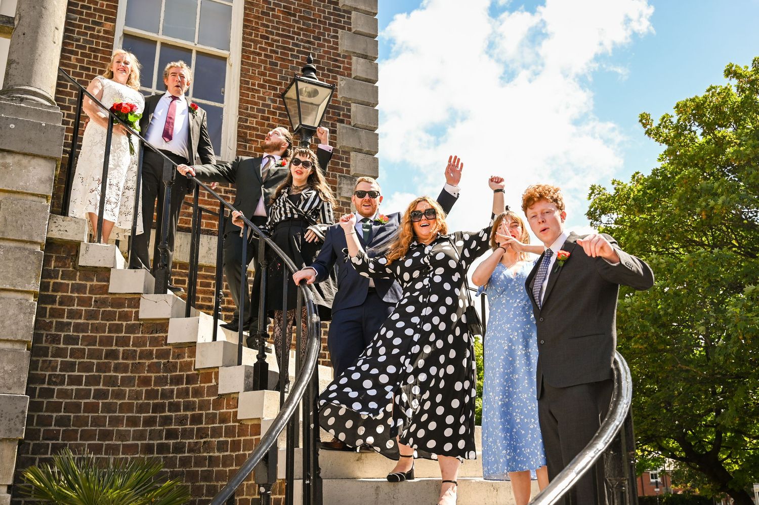 Wedding party celebrating on brick building steps, guests waving and cheering in festive attire on a sunny day.