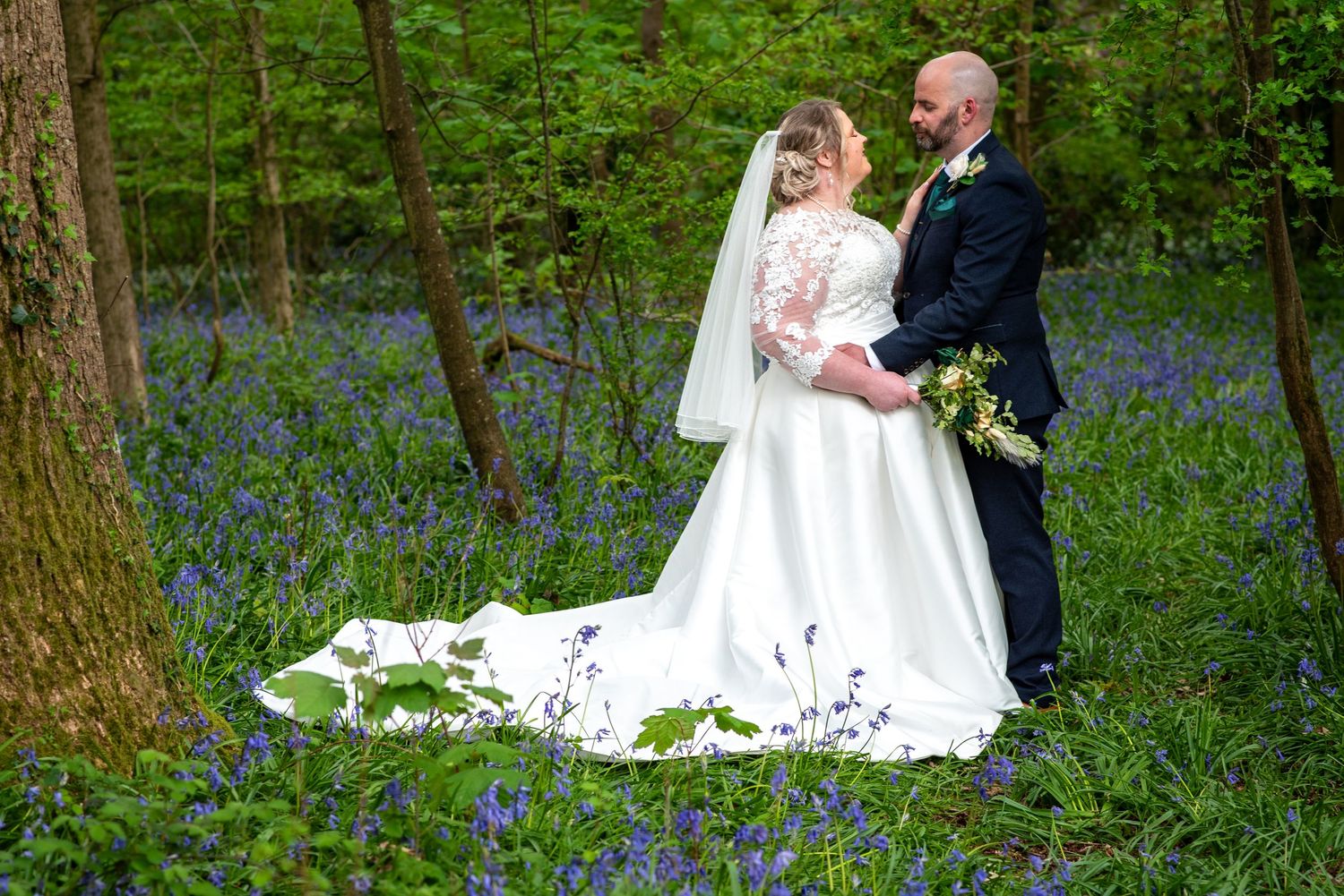 Bride and groom embrace in a bluebell woodland, bride wearing a white gown with long train and veil.