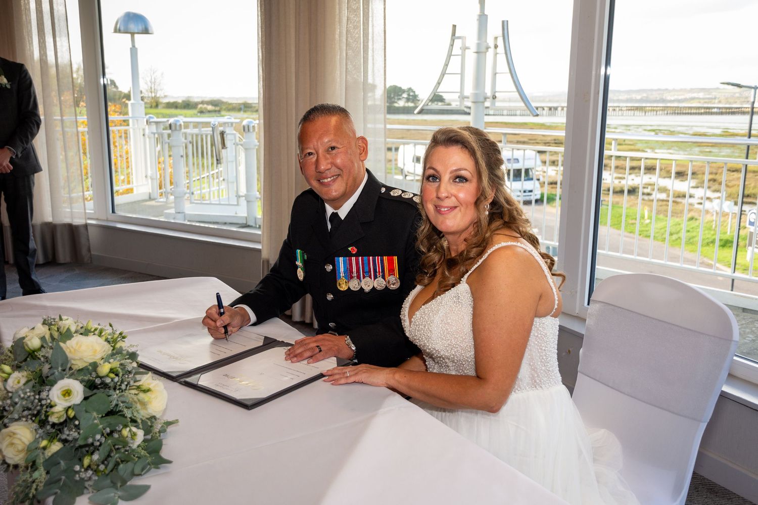 Military groom in uniform with medals signs wedding register beside smiling bride in white dress at waterfront venue.