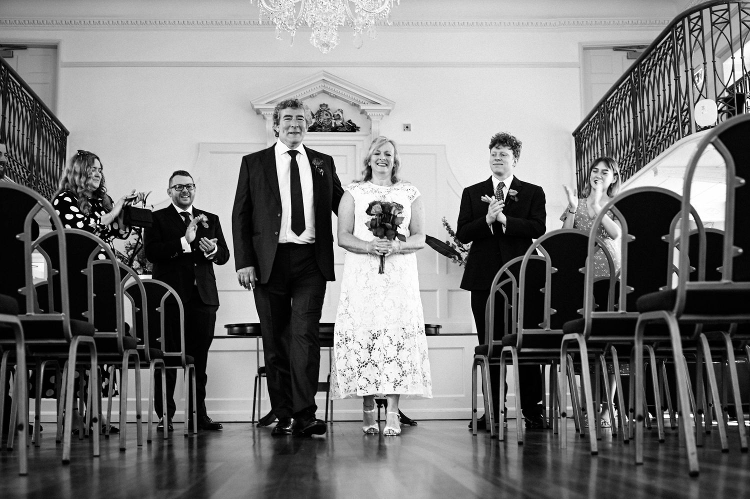 Black and white photo of a couple walking down the aisle at a wedding ceremony in an elegant hall.