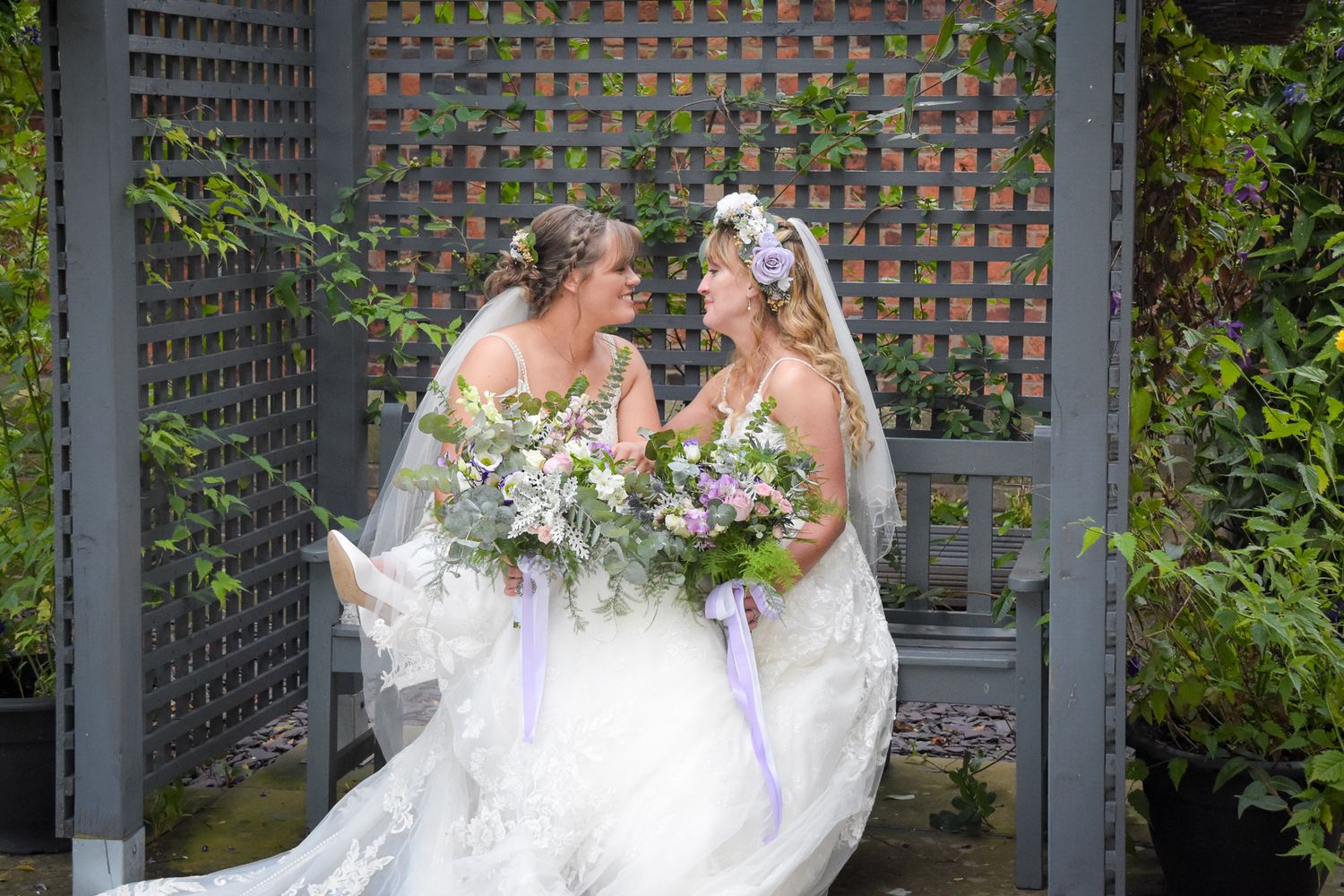 Two brides in white gowns holding floral bouquets smile at each other under a garden arbor.