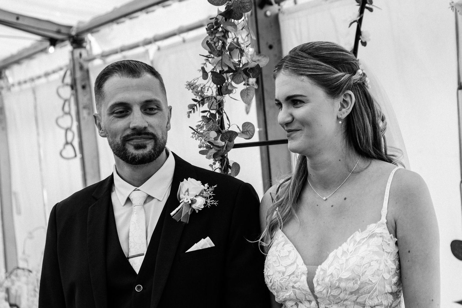 Black and white photo of a bride and groom smiling during their wedding ceremony under a floral arch.
