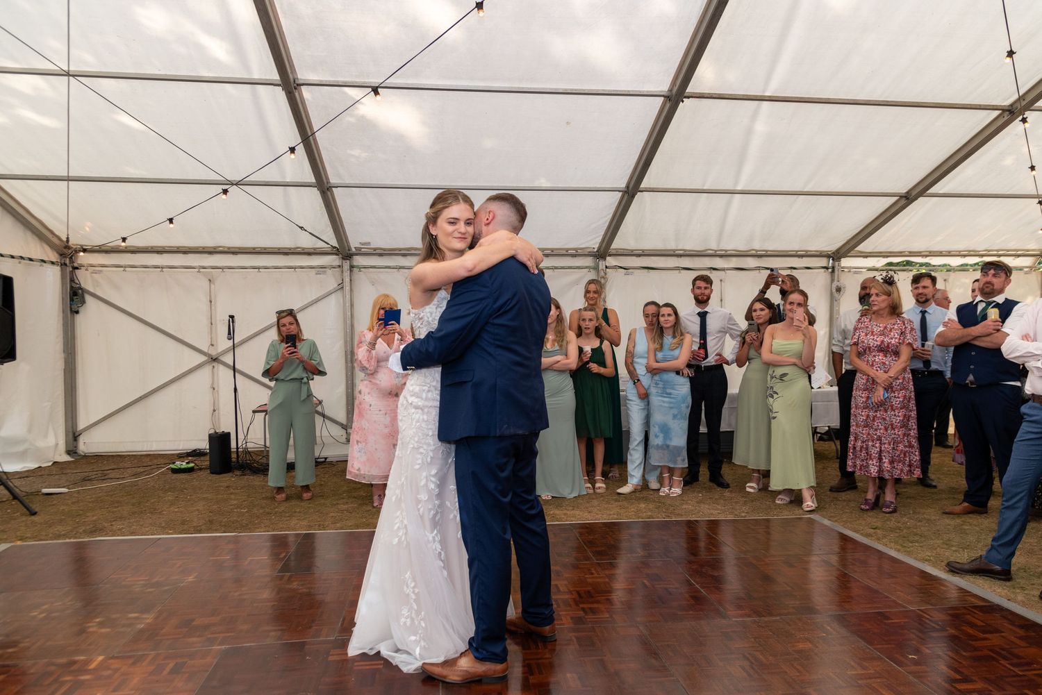 Bride and groom share first dance in marquee wedding venue with guests watching in background.