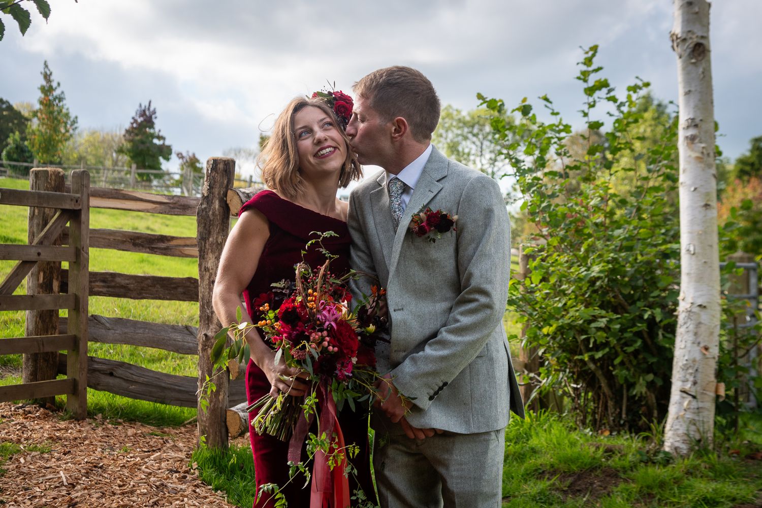 Couple in wedding attire sharing a kiss outdoors, bride in burgundy dress holding red bouquet, groom in grey suit.