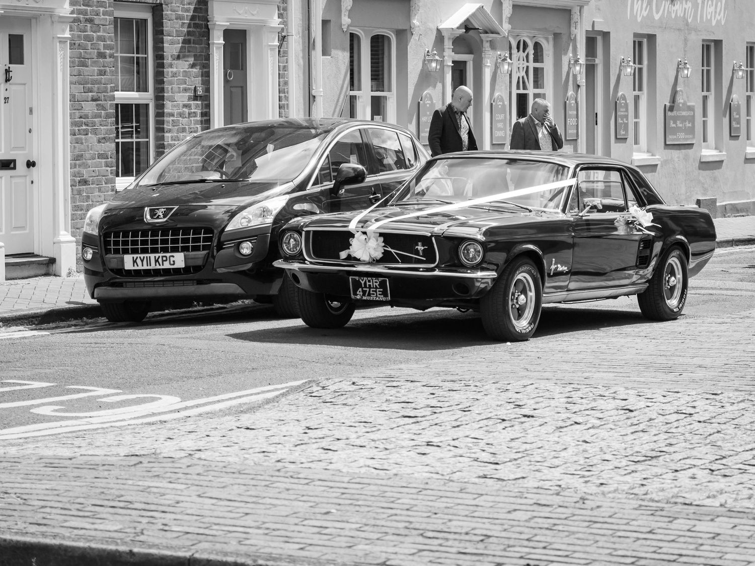 Black and white photo of a classic Ford Mustang and modern SUV parked on a cobblestone street near brick buildings.