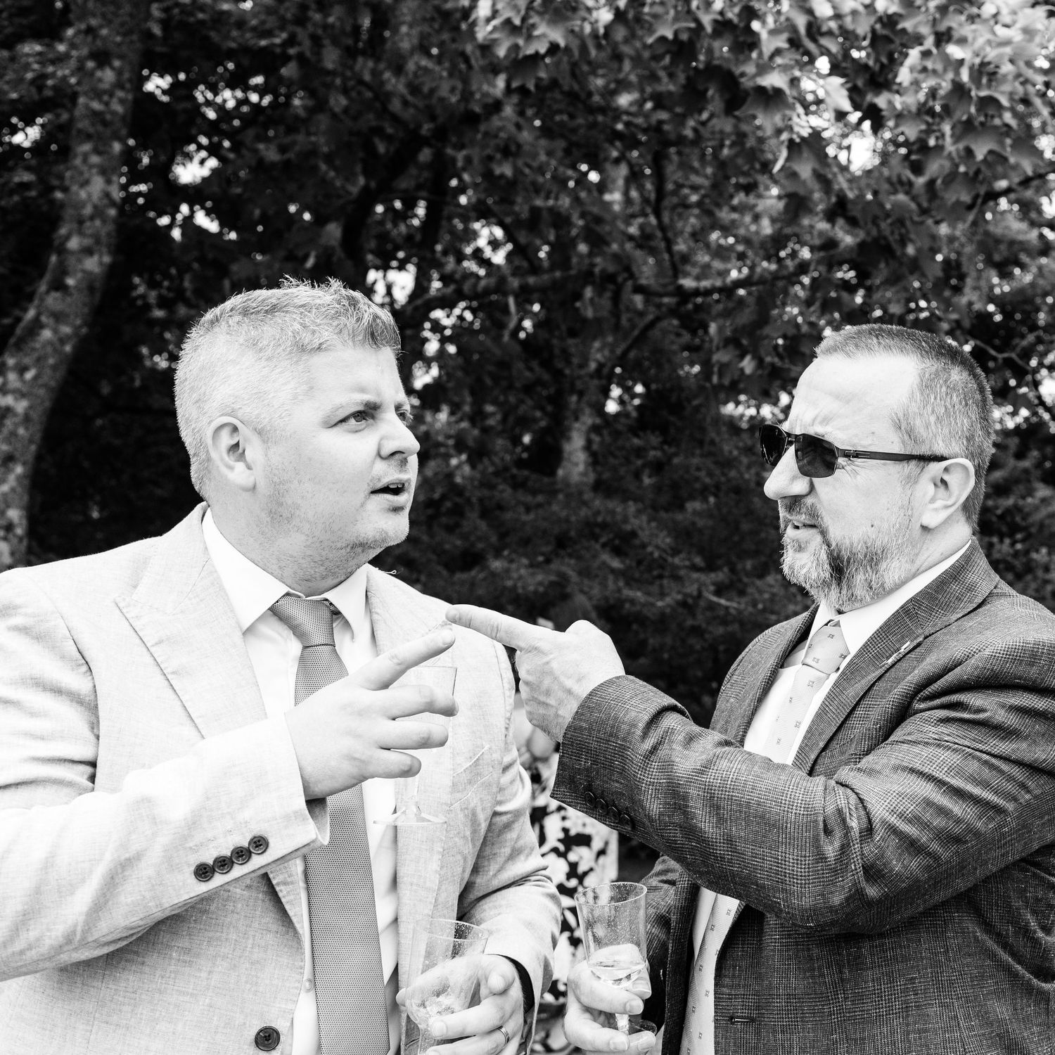 Two men in suits adjust a tie outdoors in a black and white wedding photo.