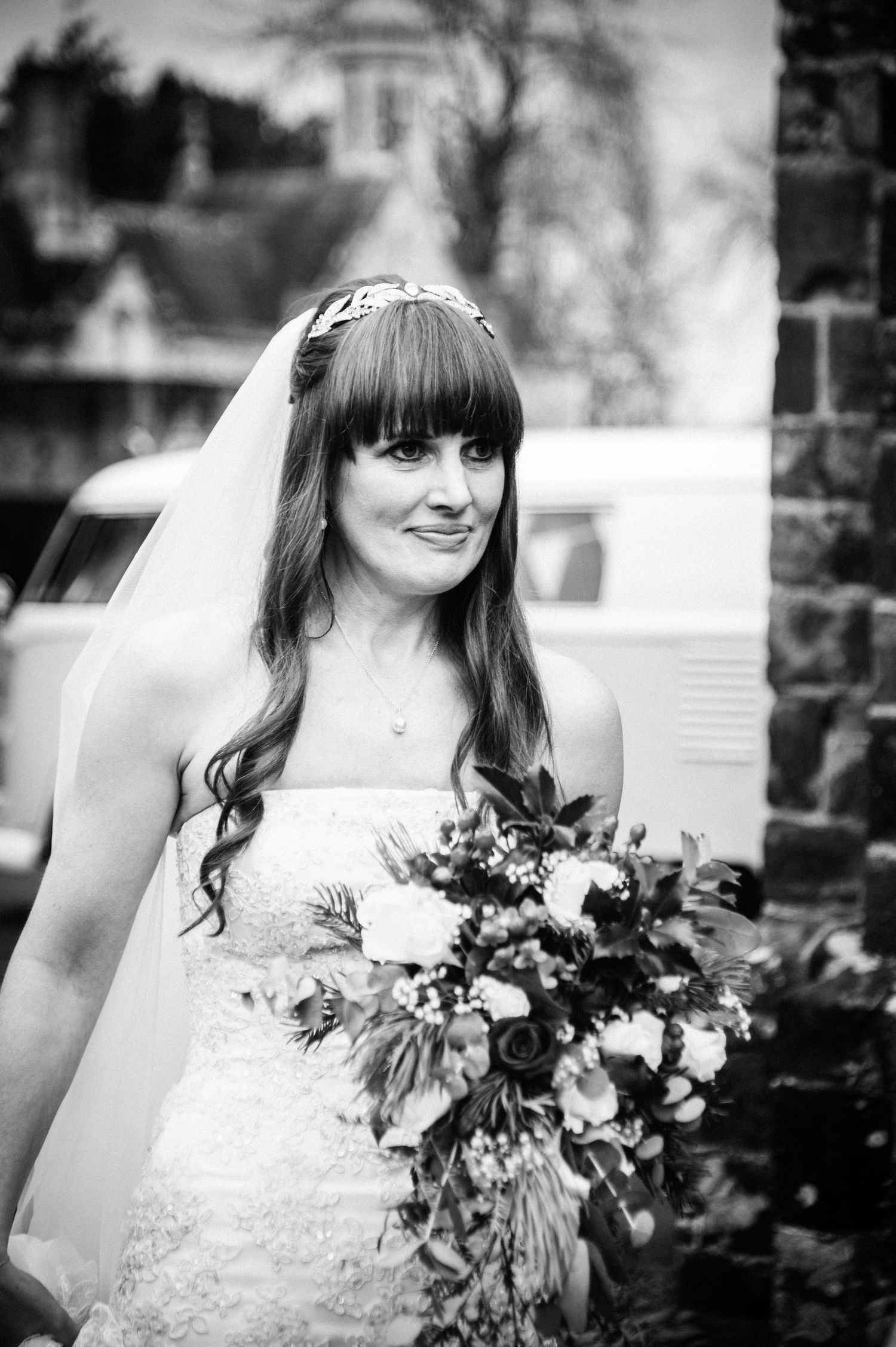 Black and white photo of a smiling bride in a strapless lace gown, holding a bouquet, wearing a veil outdoors.