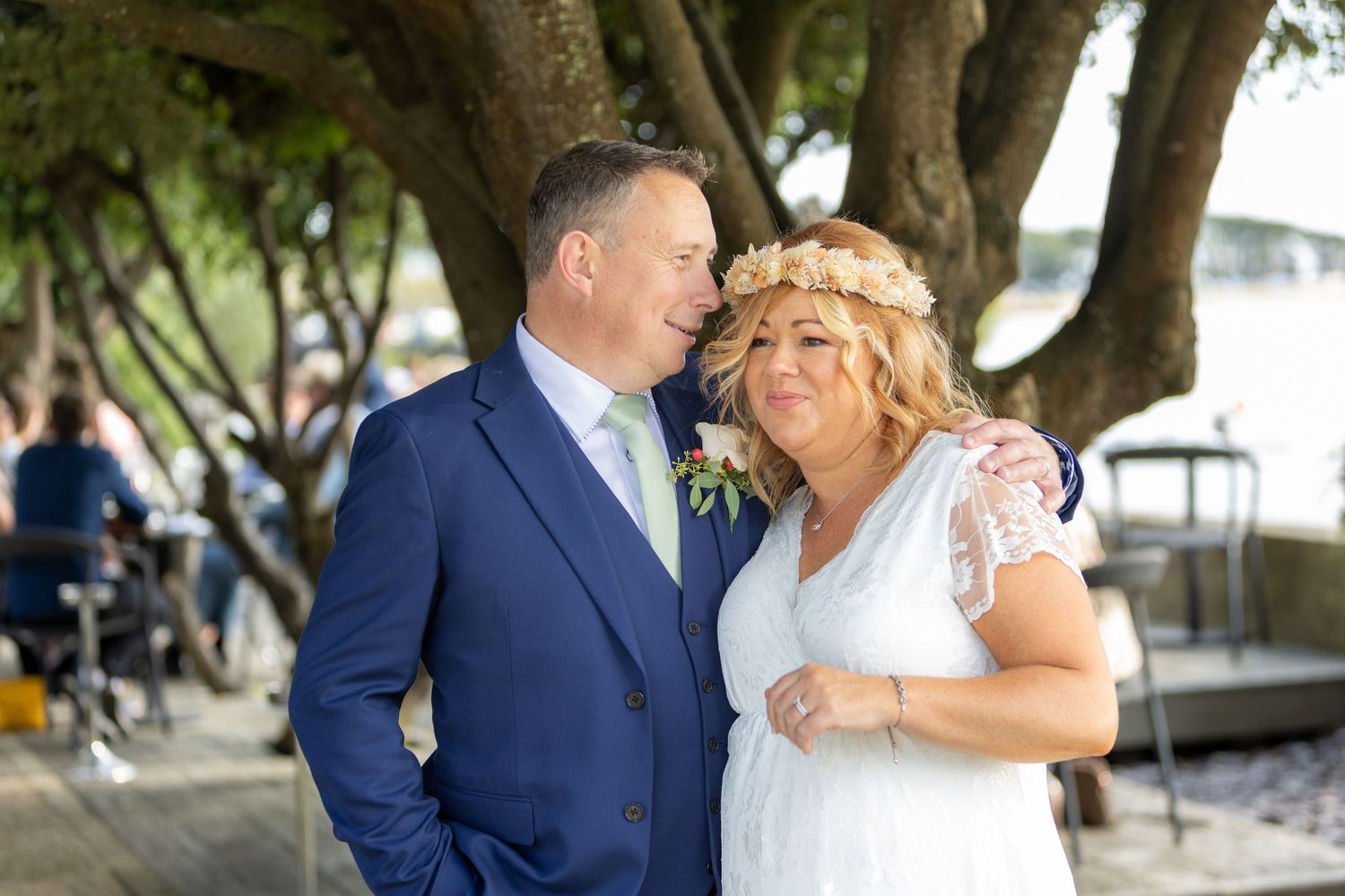 Newlywed couple shares a tender moment under a tree during their outdoor waterfront wedding ceremony.