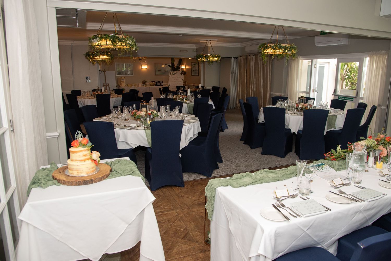 Elegant wedding reception venue with white tablecloths, navy blue chairs and crystal chandeliers in a bright banquet hall.