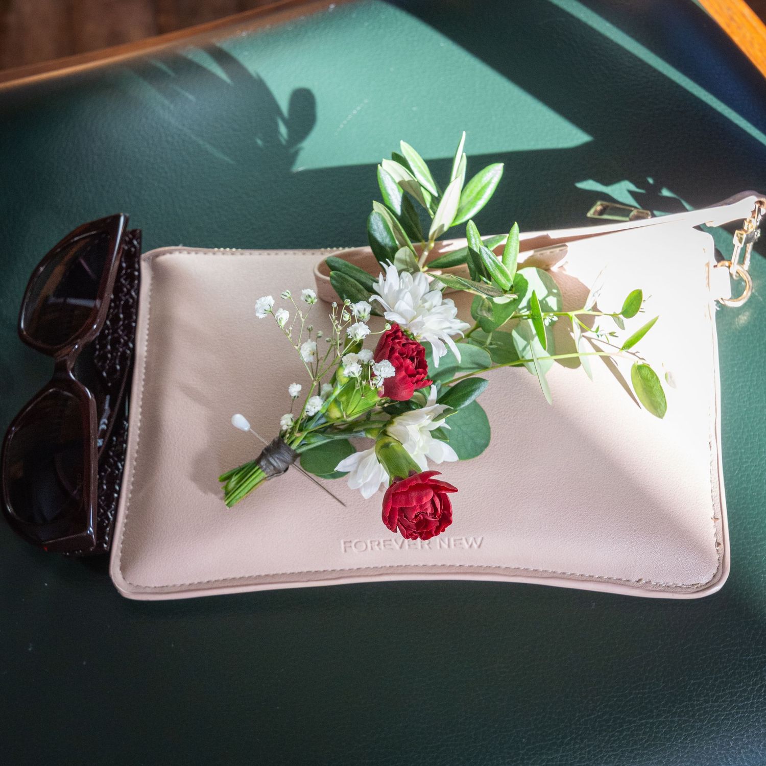 White leather clutch purse with red roses and greenery bouquet beside black sunglasses on green surface.