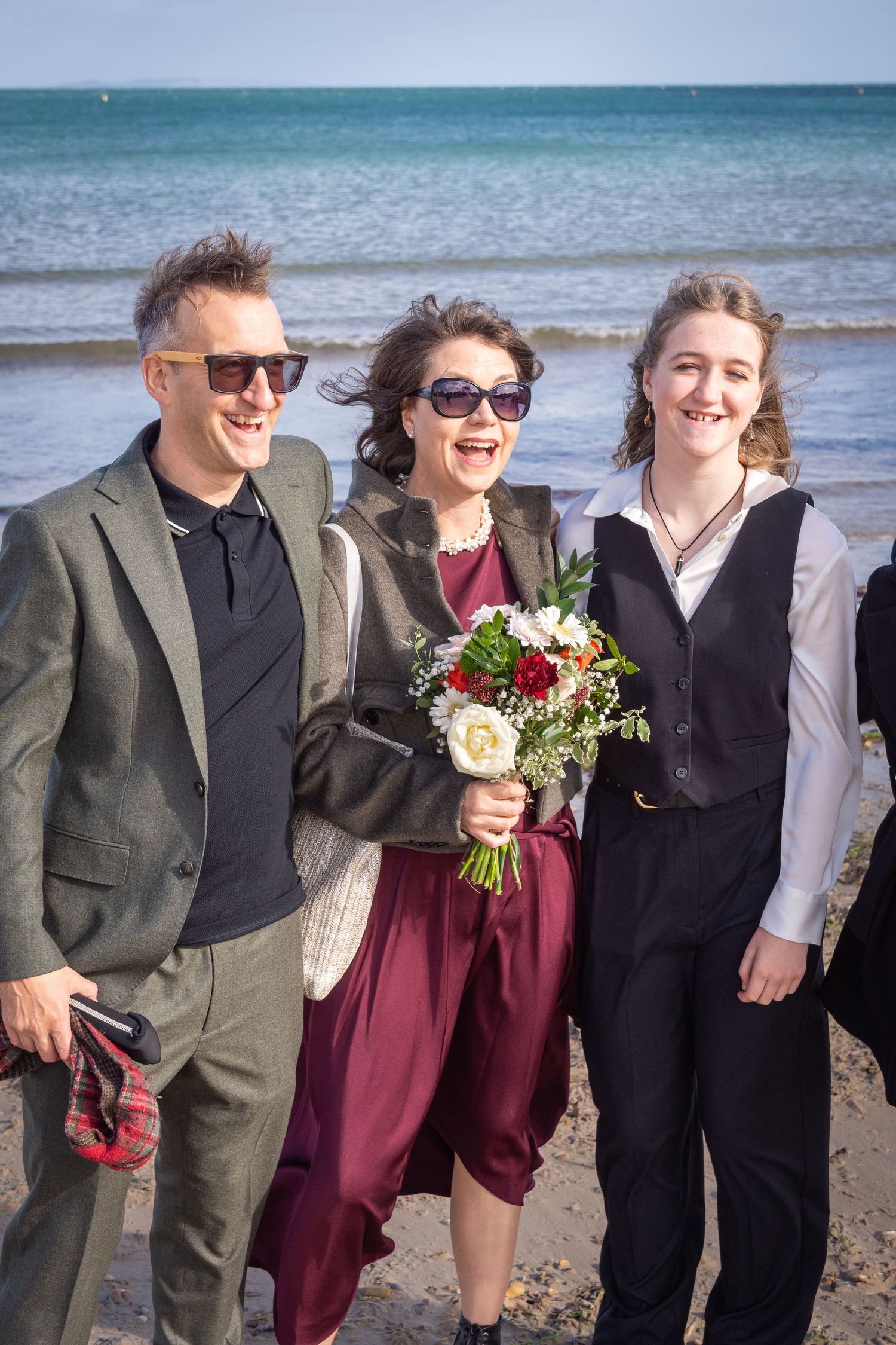 Three people smile on beach at wedding, woman holds colorful bouquet by ocean.
