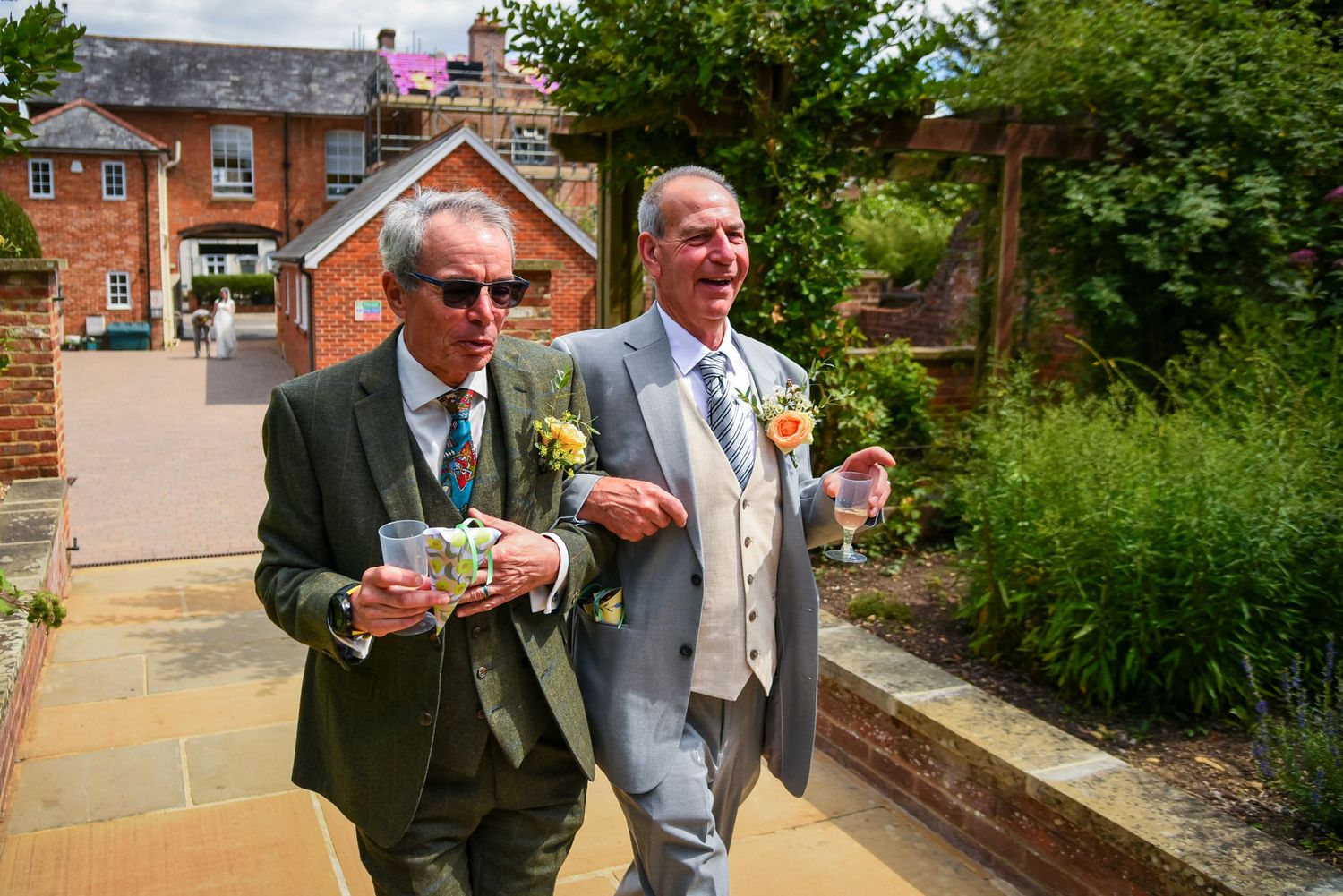 Two smartly dressed men wearing boutonnieres walking together outdoors at a wedding reception.