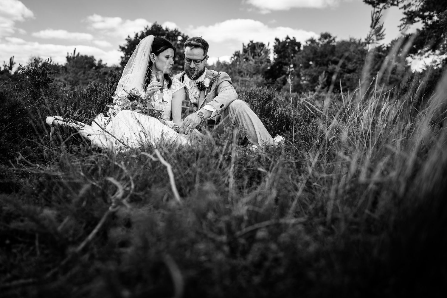 Black and white photo of a bride and groom sitting together in tall grass, sharing an intimate moment outdoors.