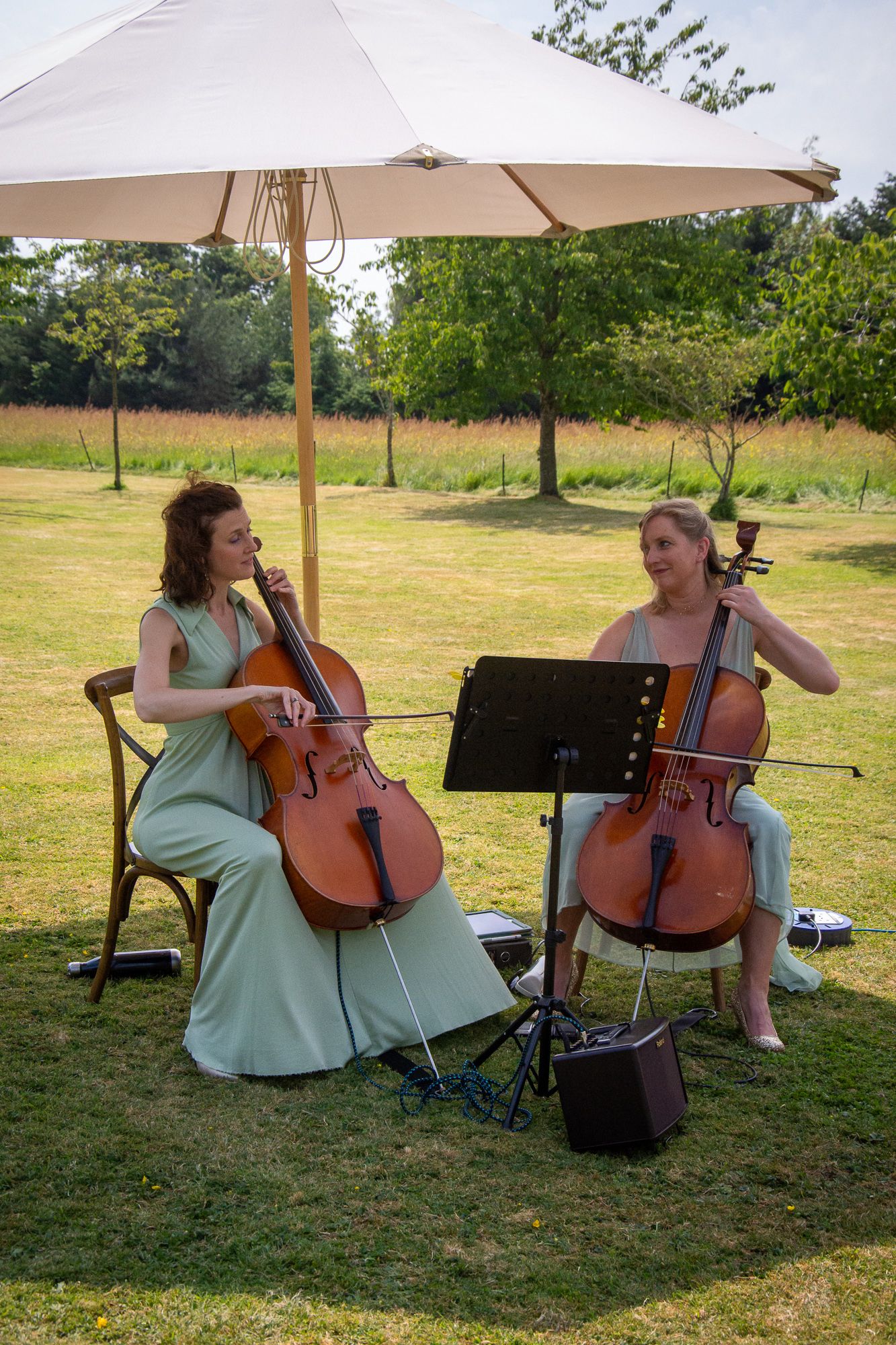 Two women in sage green dresses playing cellos outdoors under a white umbrella on a sunny day.
