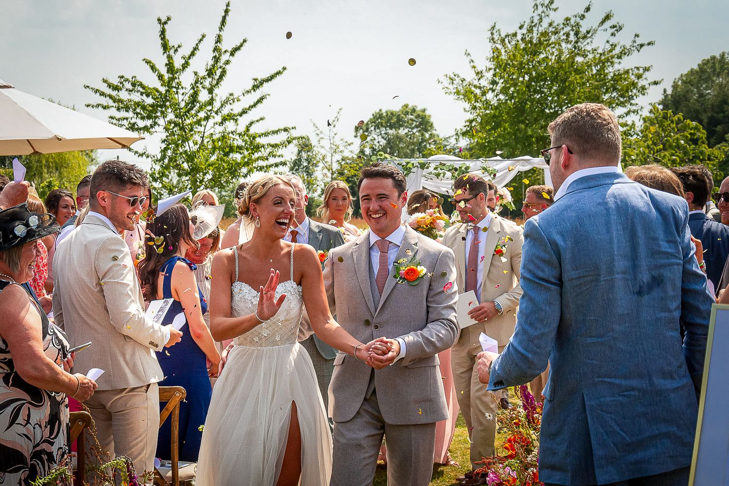 Bride and groom laughing as guests throw confetti during outdoor wedding ceremony on sunny day.