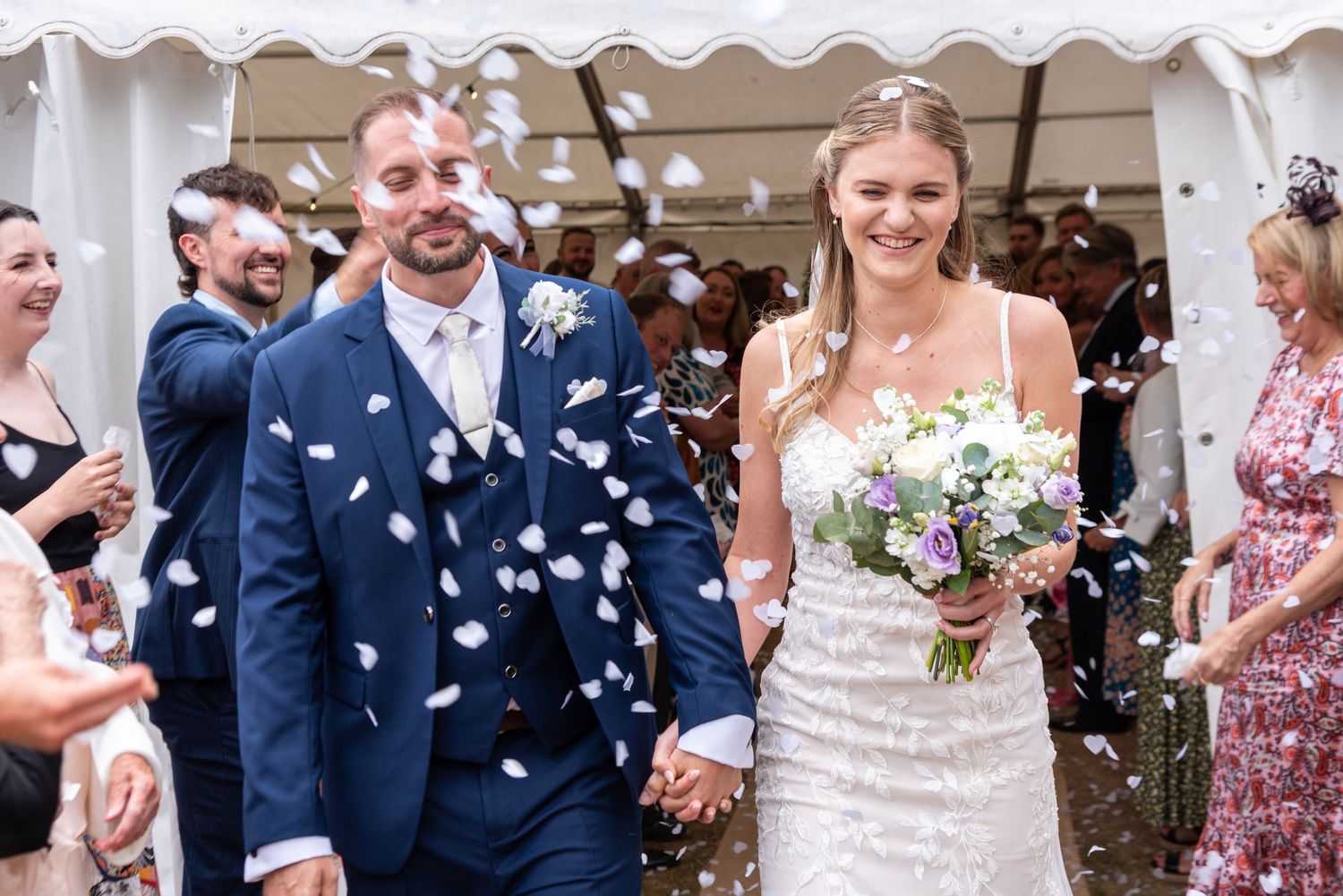 Bride and groom walk through confetti shower, guests celebrating outdoors at a sunny summer wedding ceremony.