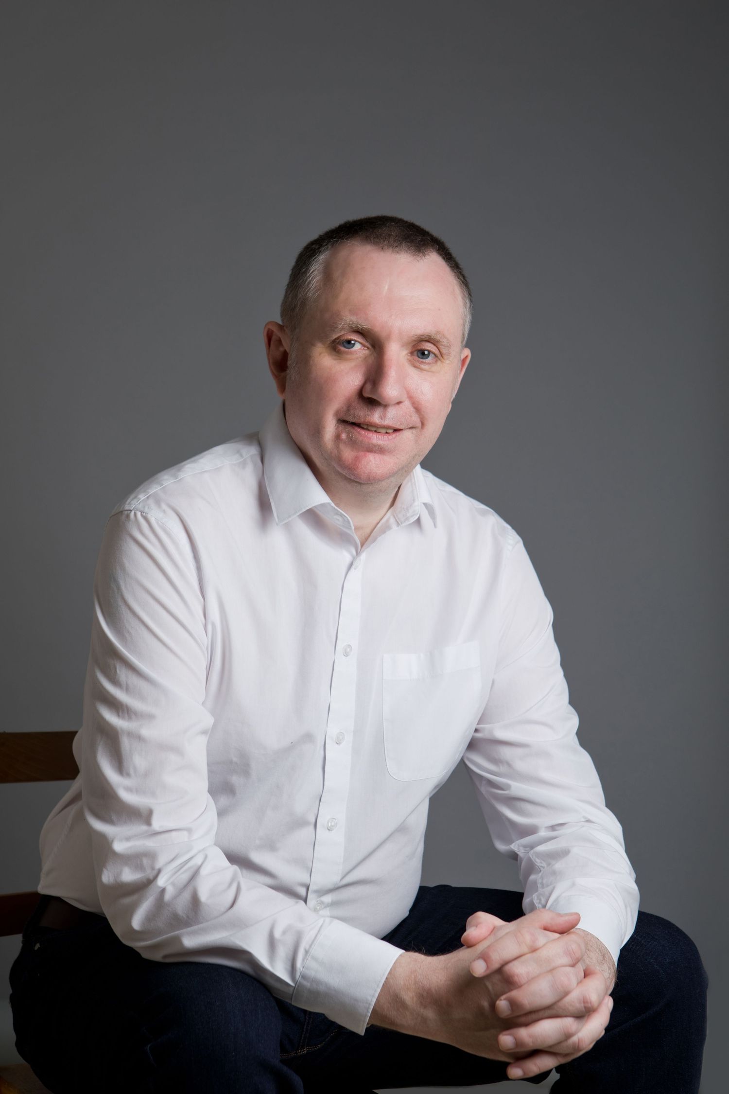 A middle-aged man in a white dress shirt sits relaxed with clasped hands, smiling against a gray background.
