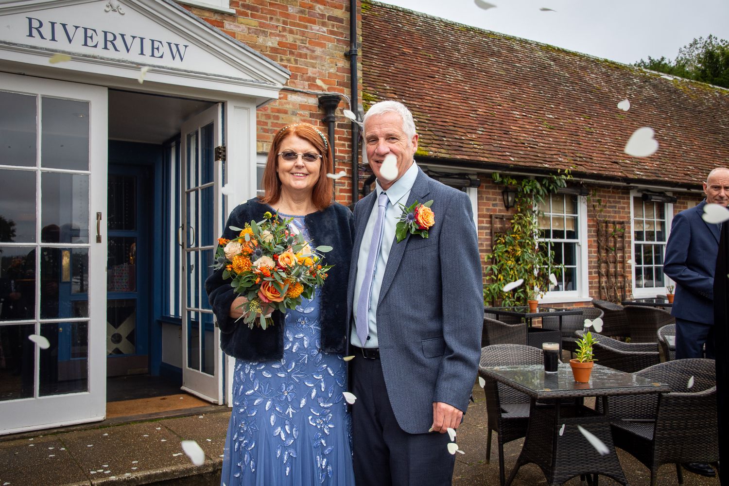 Couple posing outside Riverview venue, bride holding orange bouquet in blue floral dress, groom in gray suit with orange boutonniere.